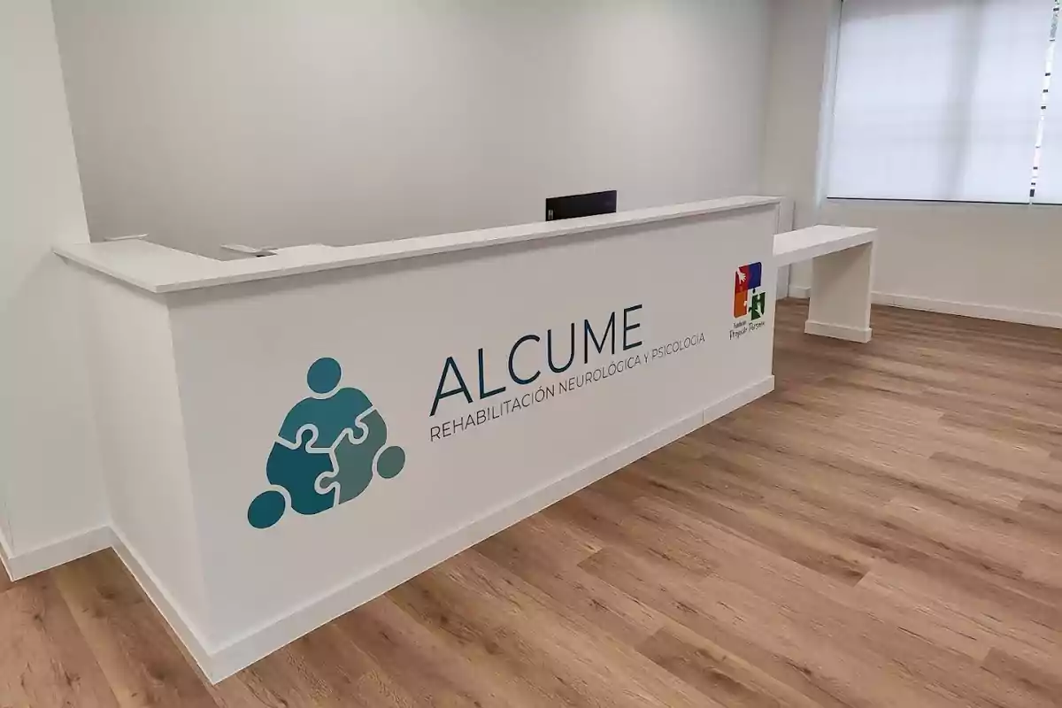 Recepción moderna de una clínica de rehabilitación neurológica y psicología llamada Alcume con su logo en el mostrador y piso de madera clara