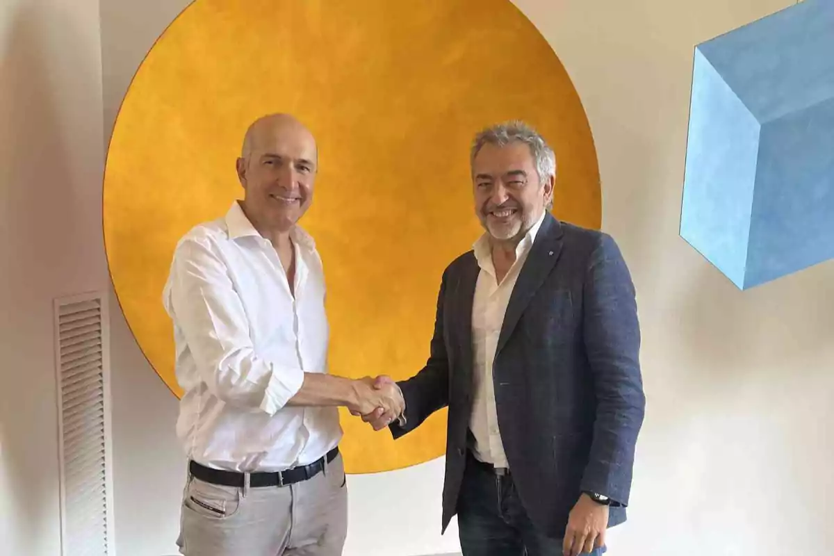 Dos hombres sonríen y se dan la mano frente a una pared con figuras geométricas de colores amarillo y azul