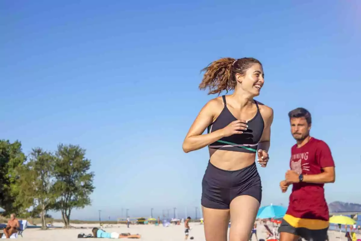 Mujer joven corriendo en la playa con ropa deportiva mientras un hombre corre detrás de ella y varias personas disfrutan del día soleado