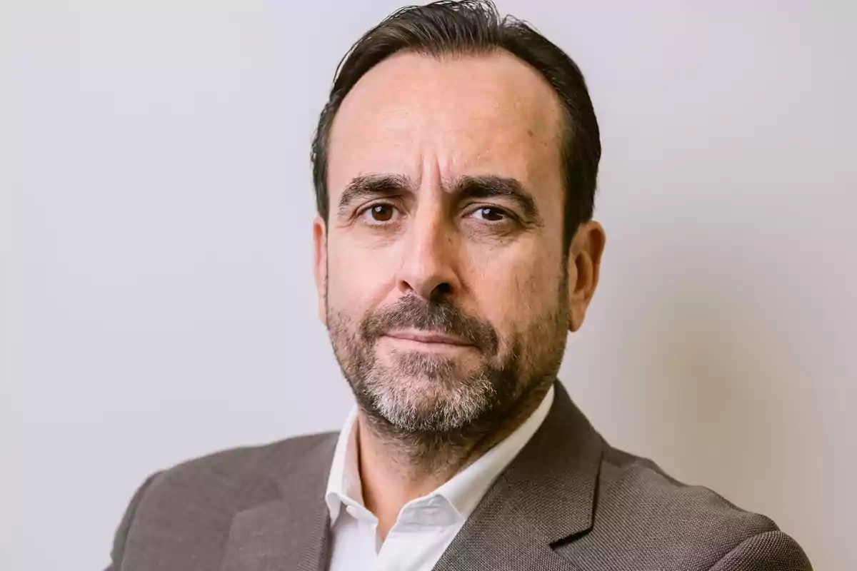 Hombre de mediana edad con barba y cabello corto vestido con saco marrón y camisa blanca mirando a la cámara con expresión seria
