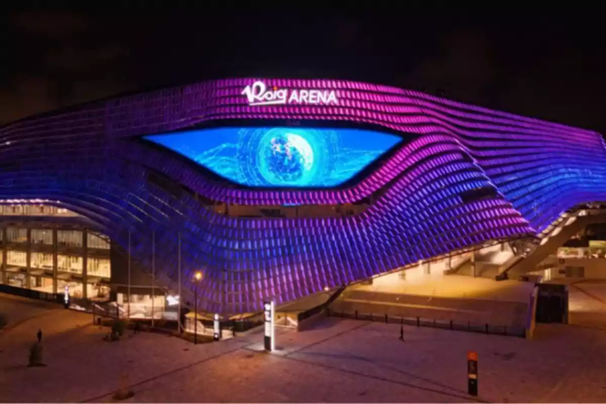 Fachada iluminada del Roig Arena en la noche con luces moradas y azules y una pantalla central que muestra un diseño futurista.