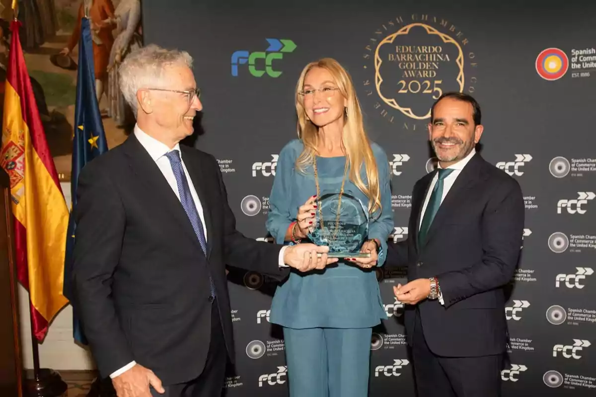 Tres personas elegantemente vestidas posan sonrientes mientras una de ellas sostiene un trofeo de cristal en un evento de premiación con banderas y un fondo institucional