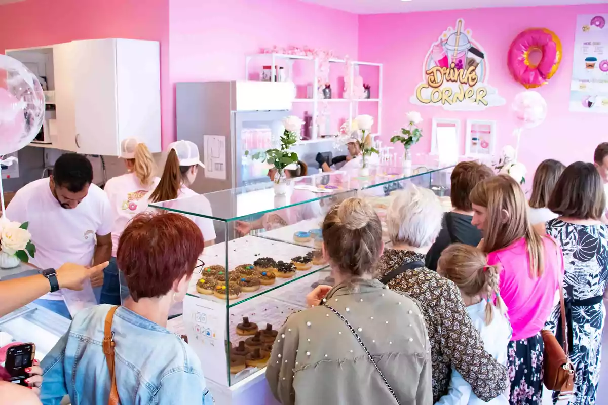 Varias personas hacen fila en una pastelería de paredes rosas mientras observan donas en una vitrina de cristal