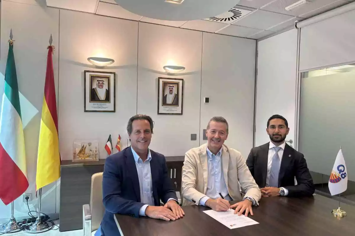 Tres hombres de negocios sentados en una mesa de oficina sonríen mientras uno de ellos firma un documento con banderas de Kuwait y España al fondo