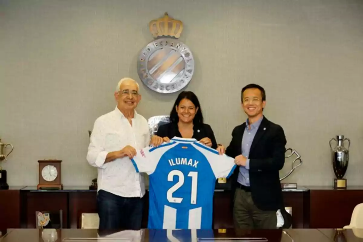 Tres personas sonríen mientras sostienen una camiseta de fútbol azul y blanca con el número 21 y la palabra ILUMAX en una oficina con trofeos y el escudo del RCD Espanyol de Barcelona al fondo