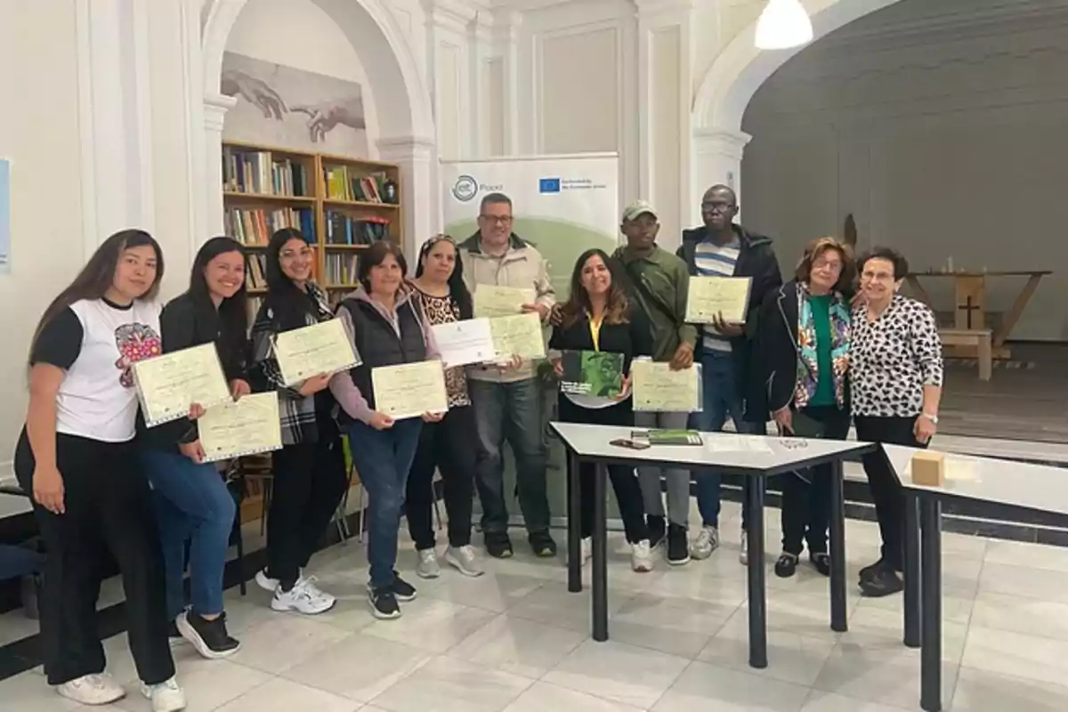 Un grupo de personas sonríe mientras sostiene diplomas en una sala con estanterías de libros y una mesa al frente
