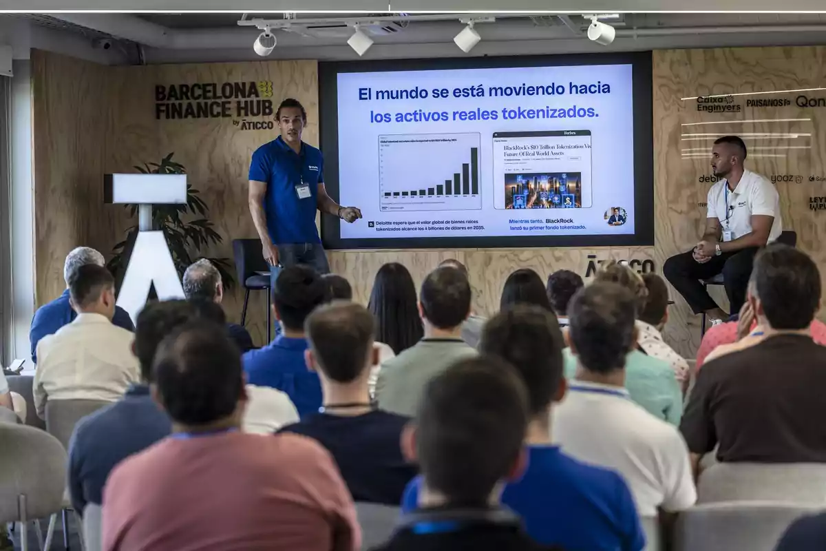 Un hombre de pie y otro sentado presentan una charla sobre activos tokenizados ante una audiencia en un evento en Barcelona Finance Hub