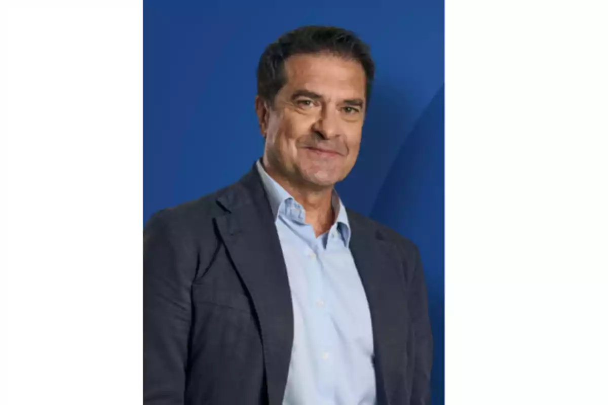 Hombre de mediana edad con cabello corto y oscuro vistiendo camisa azul clara y saco azul marino sobre fondo azul
