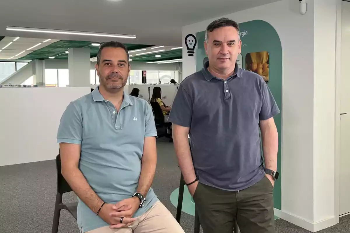 Dos hombres posan en una oficina moderna con escritorios y personas trabajando al fondo