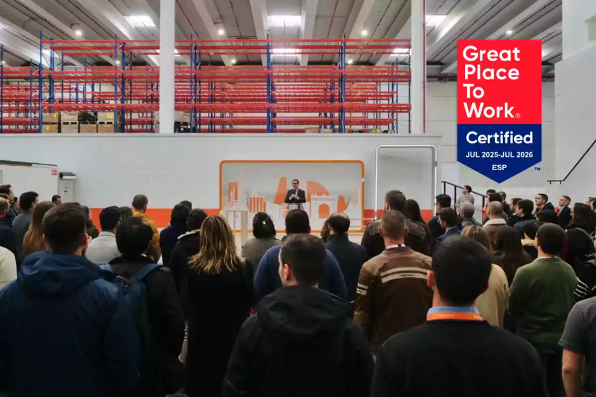 Un grupo de personas escucha a un orador en una nave industrial con estanterías vacías y un cartel de certificación Great Place to Work en la pared