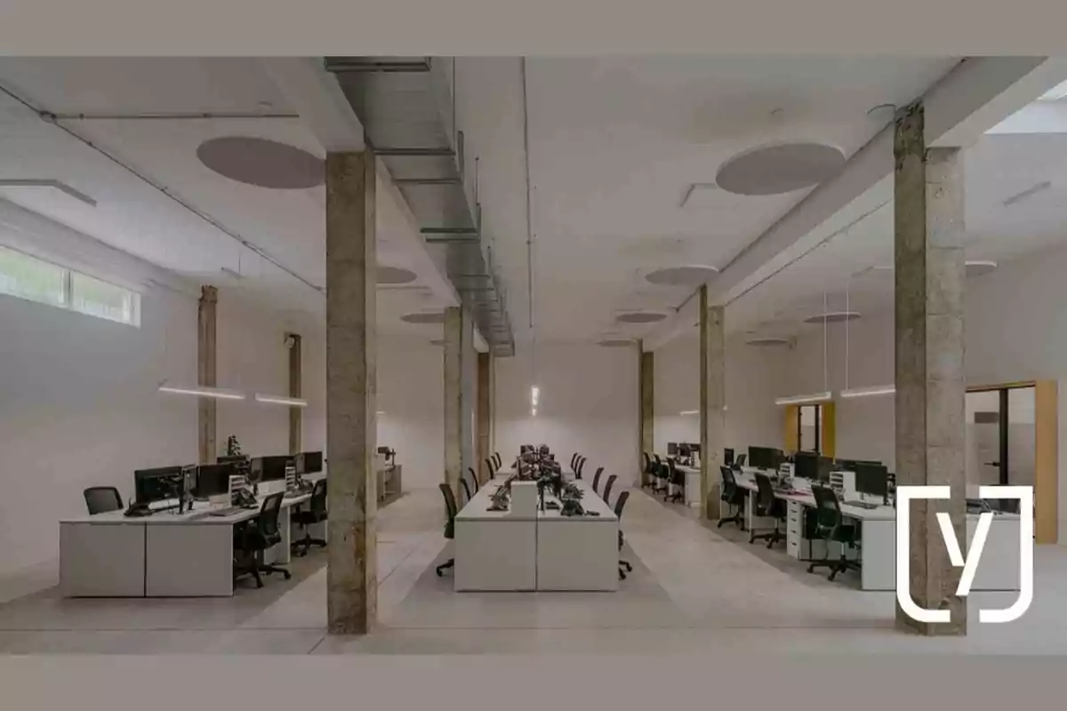 Oficina moderna con escritorios alineados, sillas negras y columnas de concreto en un espacio amplio y bien iluminado