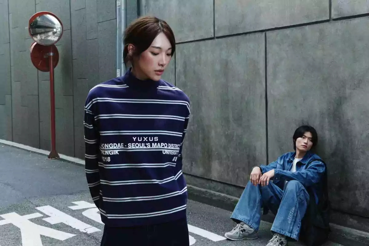 Dos personas en una calle urbana, una de pie con suéter de rayas y otra sentada en el suelo junto a una pared de concreto
