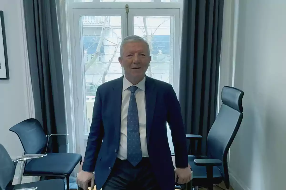 Hombre de cabello canoso con traje azul y corbata de pie en una oficina moderna frente a una ventana grande Hombre de cabello canoso con traje azul y corbata de pie en una oficina moderna frente a una ventana grande