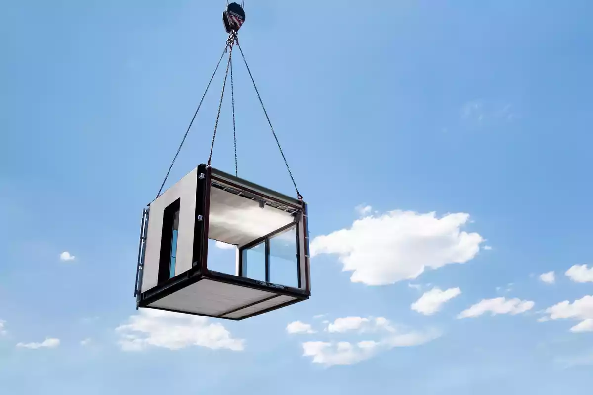 Un módulo prefabricado de construcción es elevado por una grúa sobre un cielo azul con algunas nubes