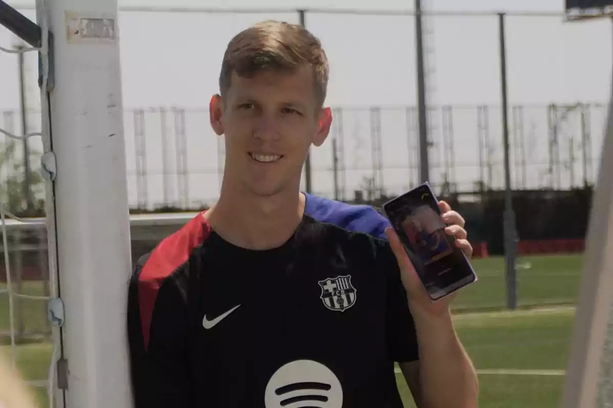 Joven con camiseta del FC Barcelona sonríe y muestra un teléfono móvil en un campo de fútbol