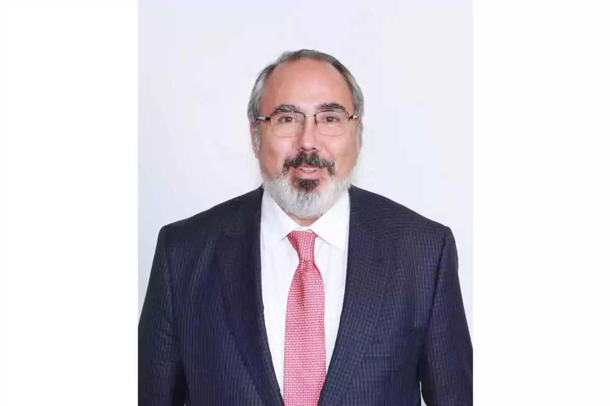 Hombre de mediana edad con barba y gafas vistiendo traje oscuro, camisa blanca y corbata roja posando frente a un fondo blanco