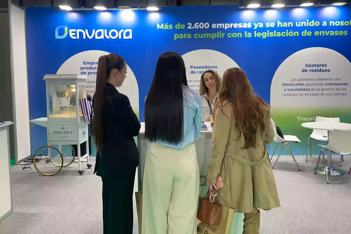 Tres mujeres conversan con una expositora en un stand de Envalora durante una feria, con una máquina de palomitas y carteles informativos de fondo.