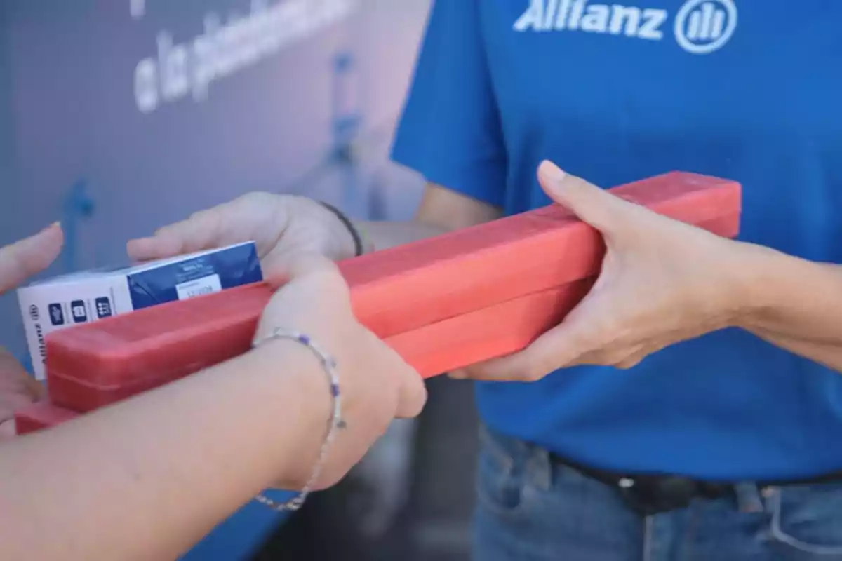 Persona con camiseta azul entrega dos objetos rojos rectangulares y una caja pequeña a otra persona