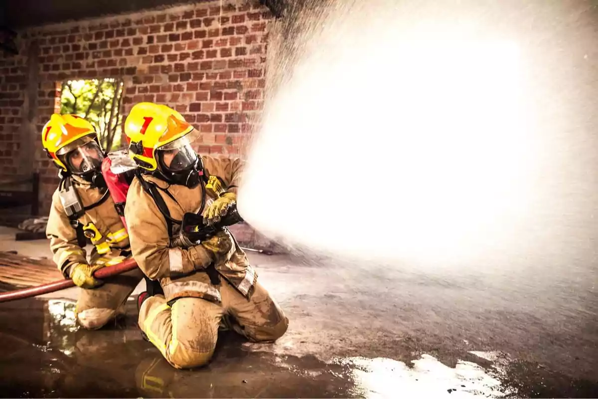 Dos bomberos con equipo de protección y cascos amarillos utilizan una manguera para rociar agua dentro de un edificio de ladrillo