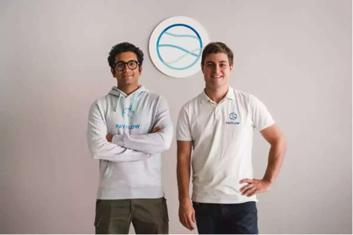 Dos personas posan sonrientes frente a una pared clara con el logo de Payflow, una lleva sudadera gris y la otra polo blanco, ambos con el mismo logotipo.