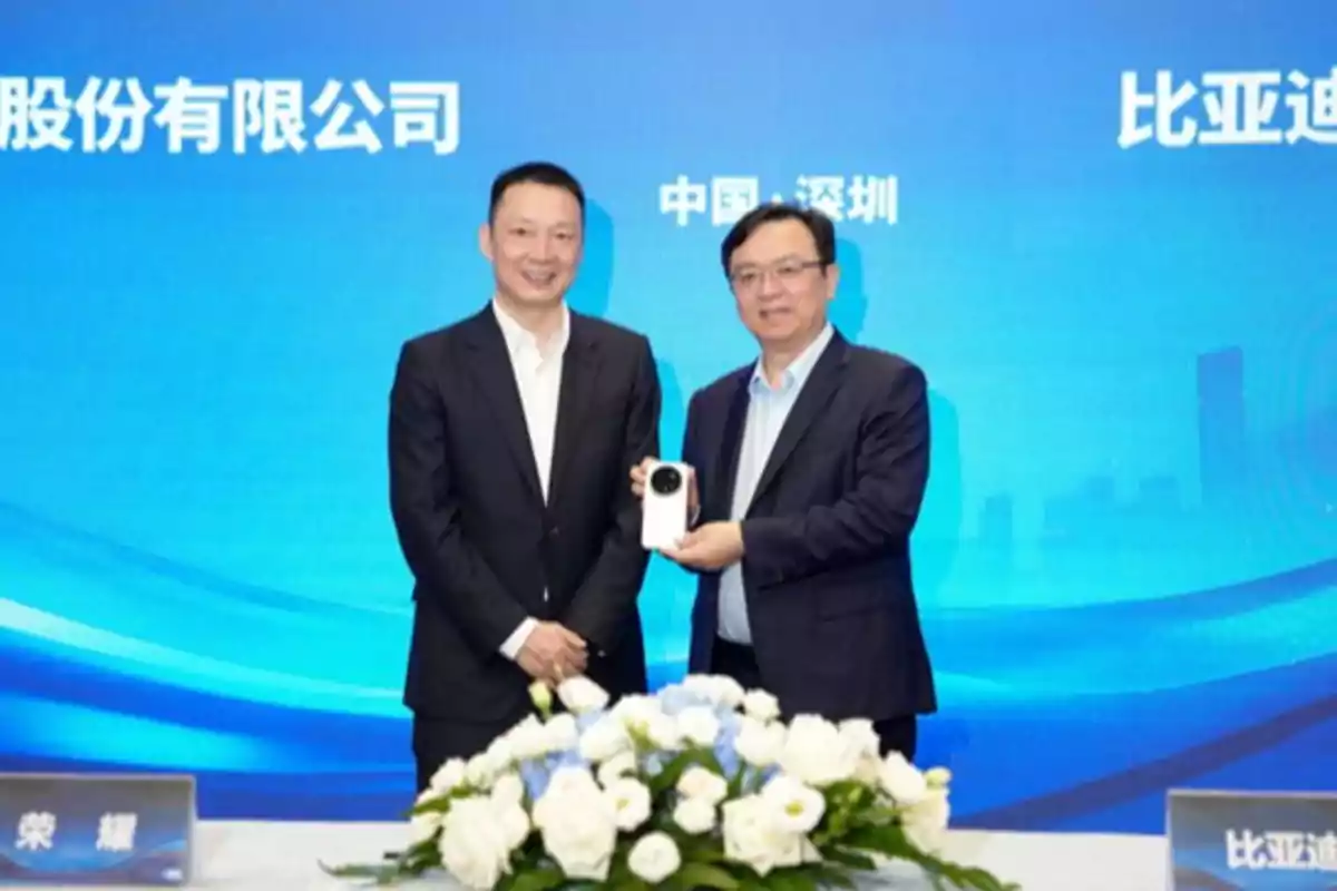 Dos hombres de traje posan sonrientes frente a un fondo azul con letras chinas, uno de ellos sostiene un teléfono móvil blanco y en primer plano hay un arreglo de flores blancas y azules. Dos hombres de traje posan sonrientes frente a un fondo azul con letras chinas, uno de ellos sostiene un teléfono móvil blanco y en primer plano hay un arreglo de flores blancas y azules.