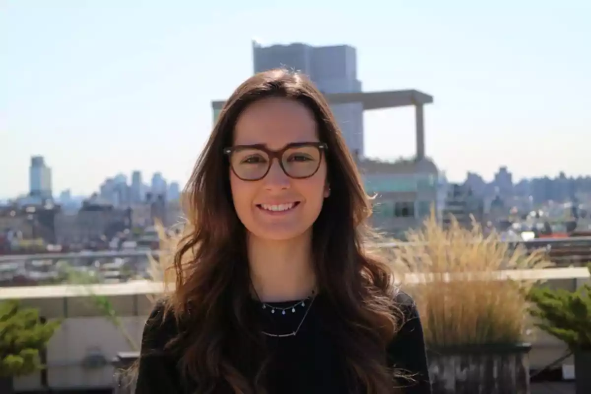 Mujer joven con gafas y cabello largo sonríe al aire libre con una ciudad de fondo borroso