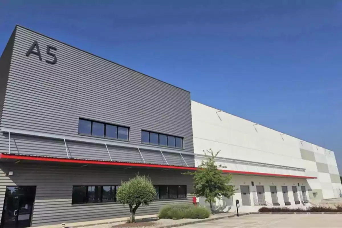 Fachada de una nave industrial moderna con paredes grises y blancas, ventanas rectangulares, puertas de carga y un letrero grande con la inscripción A5 Fachada de una nave industrial moderna con paredes grises y blancas, ventanas rectangulares, puertas de carga y un letrero grande con la inscripción A5