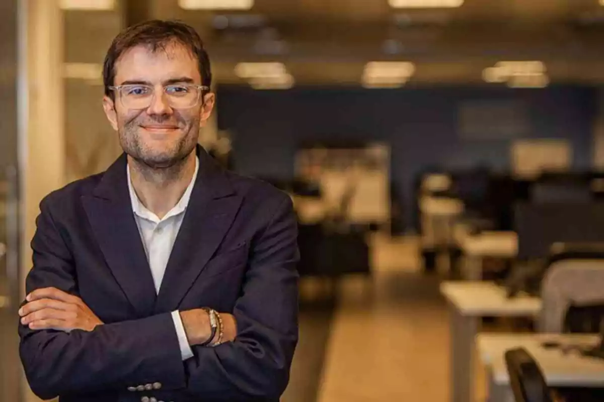 Hombre con gafas y saco azul sonriendo con los brazos cruzados en una oficina moderna desenfocada al fondo