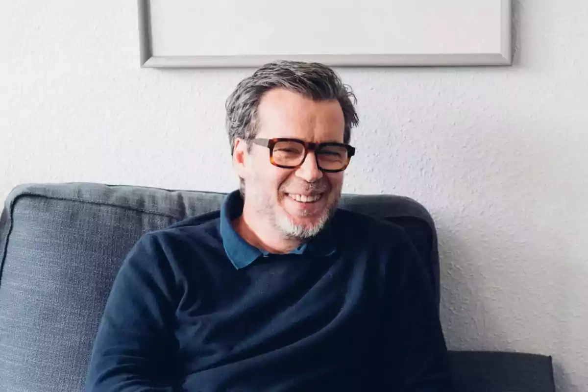 Hombre de mediana edad con gafas y barba sonriendo mientras está sentado en un sofá gris
