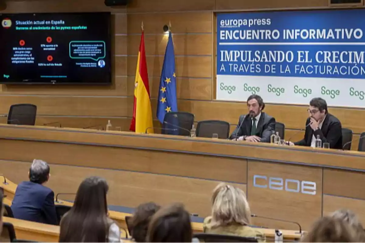 Dos hombres sentados en una mesa de conferencias durante un evento informativo con público asistente, banderas de España y la Unión Europea al fondo y una pantalla con información sobre la situación de las pymes en España. Dos hombres sentados en una mesa de conferencias durante un evento informativo con público asistente, banderas de España y la Unión Europea al fondo y una pantalla con información sobre la situación de las pymes en España.