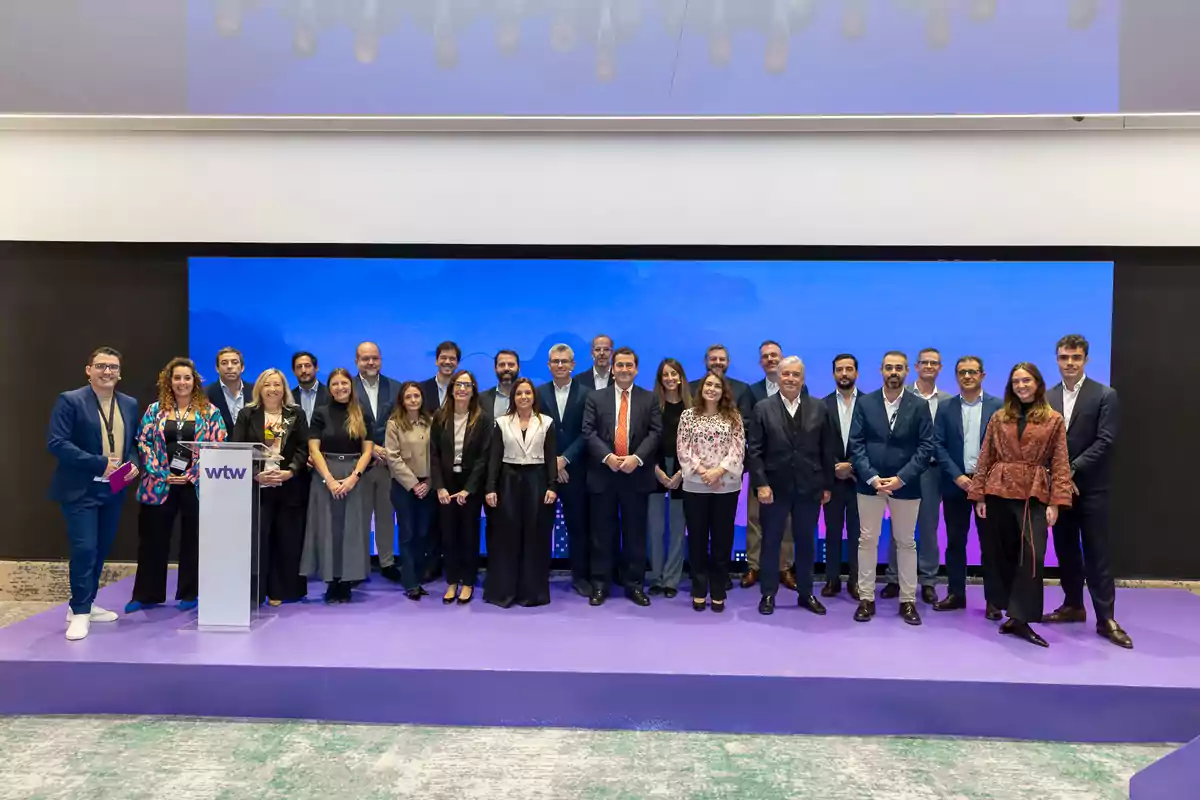 Grupo de personas posando juntas sobre un escenario con fondo azul y púrpura en un evento corporativo Grupo de personas posando juntas sobre un escenario con fondo azul y púrpura en un evento corporativo