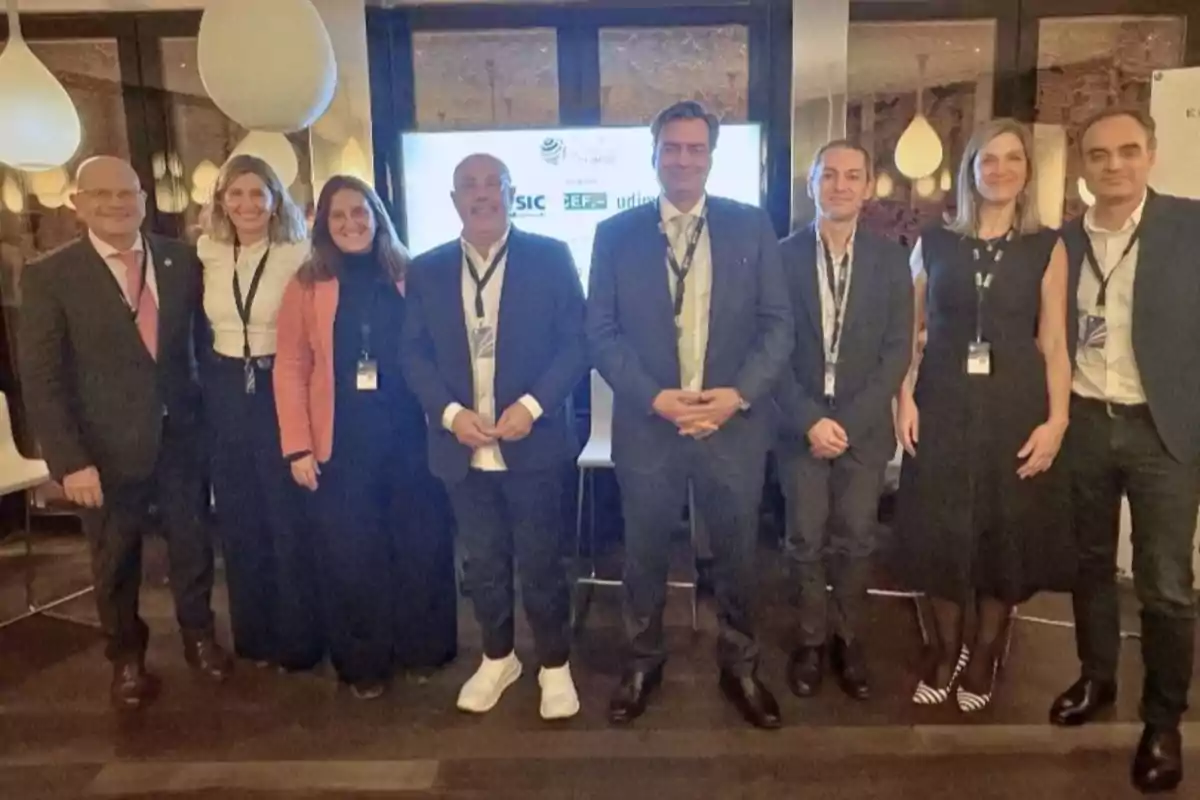 Ocho personas posan juntas en un evento formal, todas llevan gafetes y están de pie frente a una pantalla en un ambiente interior elegante.