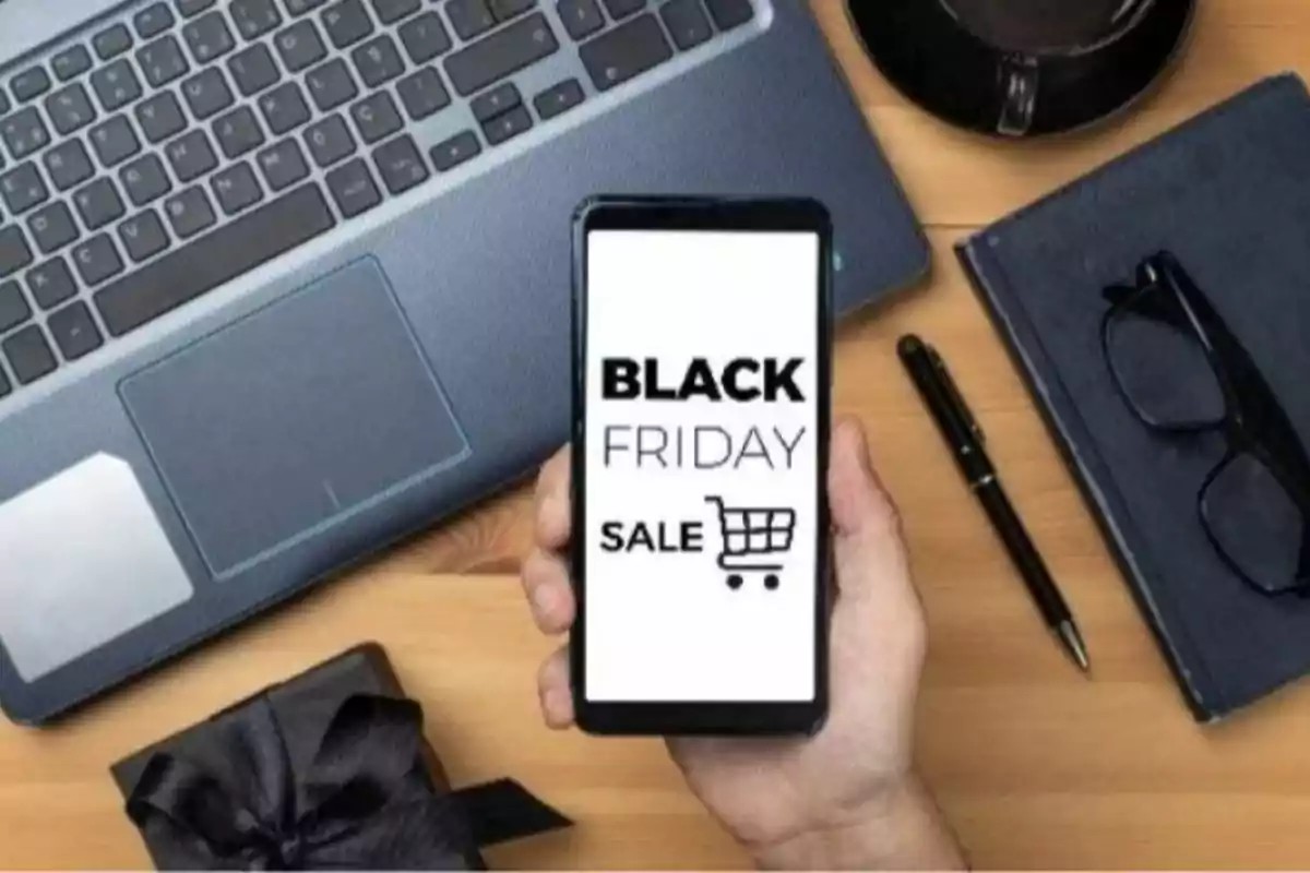 Persona sosteniendo un teléfono móvil que muestra una promoción de Black Friday junto a una laptop, una taza de café, un cuaderno, unos lentes, un bolígrafo y una caja de regalo sobre una mesa de madera