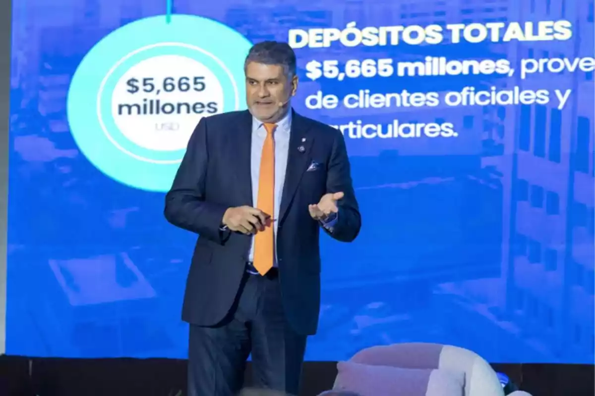 Hombre de traje oscuro y corbata naranja hablando en una presentación financiera con una pantalla azul de fondo que muestra cifras de depósitos totales por 5,665 millones de dólares