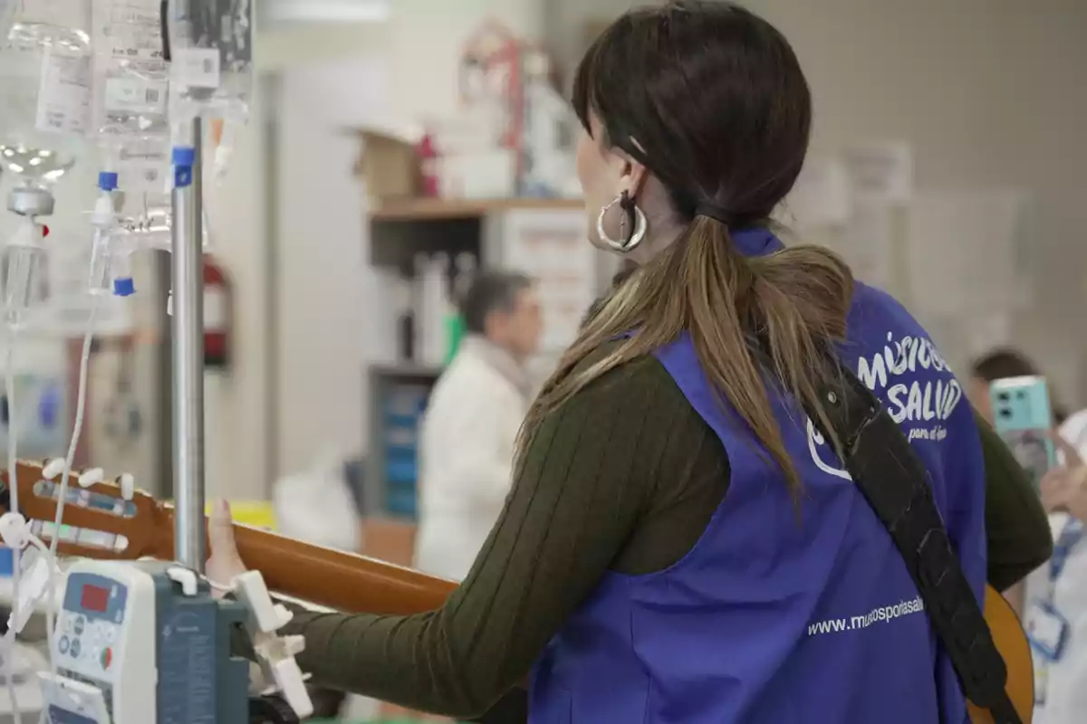 Mujer con chaleco azul tocando la guitarra en una sala de hospital junto a equipos médicos