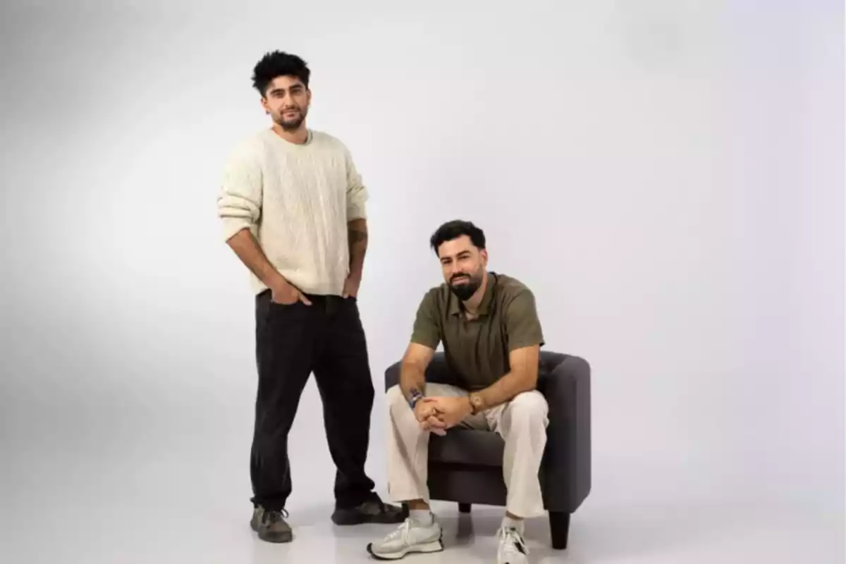 Dos hombres posan en un estudio con fondo claro uno está de pie con las manos en los bolsillos y el otro está sentado en un sillón gris