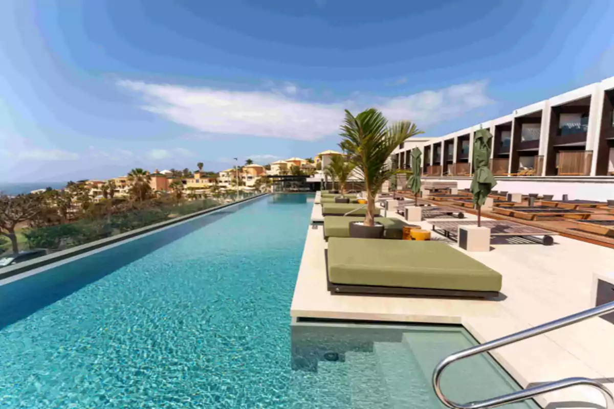 Piscina infinita con tumbonas verdes y palmeras junto a un hotel moderno bajo un cielo azul, con vistas a una zona residencial y montañosa al fondo Piscina infinita con tumbonas verdes y palmeras junto a un hotel moderno bajo un cielo azul, con vistas a una zona residencial y montañosa al fondo