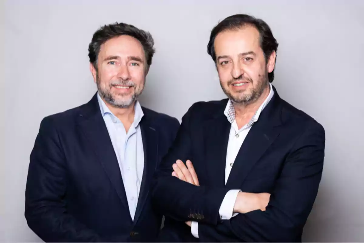 Dos hombres de mediana edad con barba y cabello oscuro posan juntos usando trajes oscuros y camisas claras frente a un fondo gris Dos hombres de mediana edad con barba y cabello oscuro posan juntos usando trajes oscuros y camisas claras frente a un fondo gris