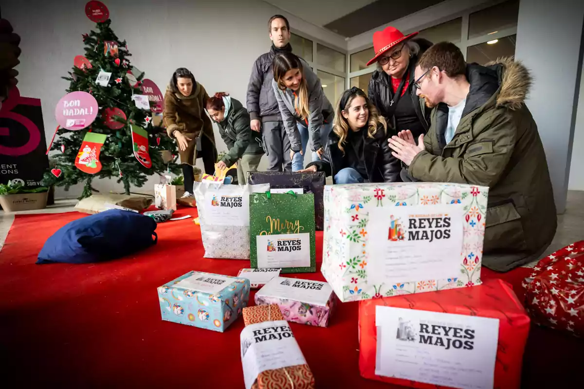 Varias personas sonríen y organizan regalos envueltos junto a un árbol de Navidad decorado, sobre una alfombra roja, con cajas que tienen etiquetas de Reyes Majos.
