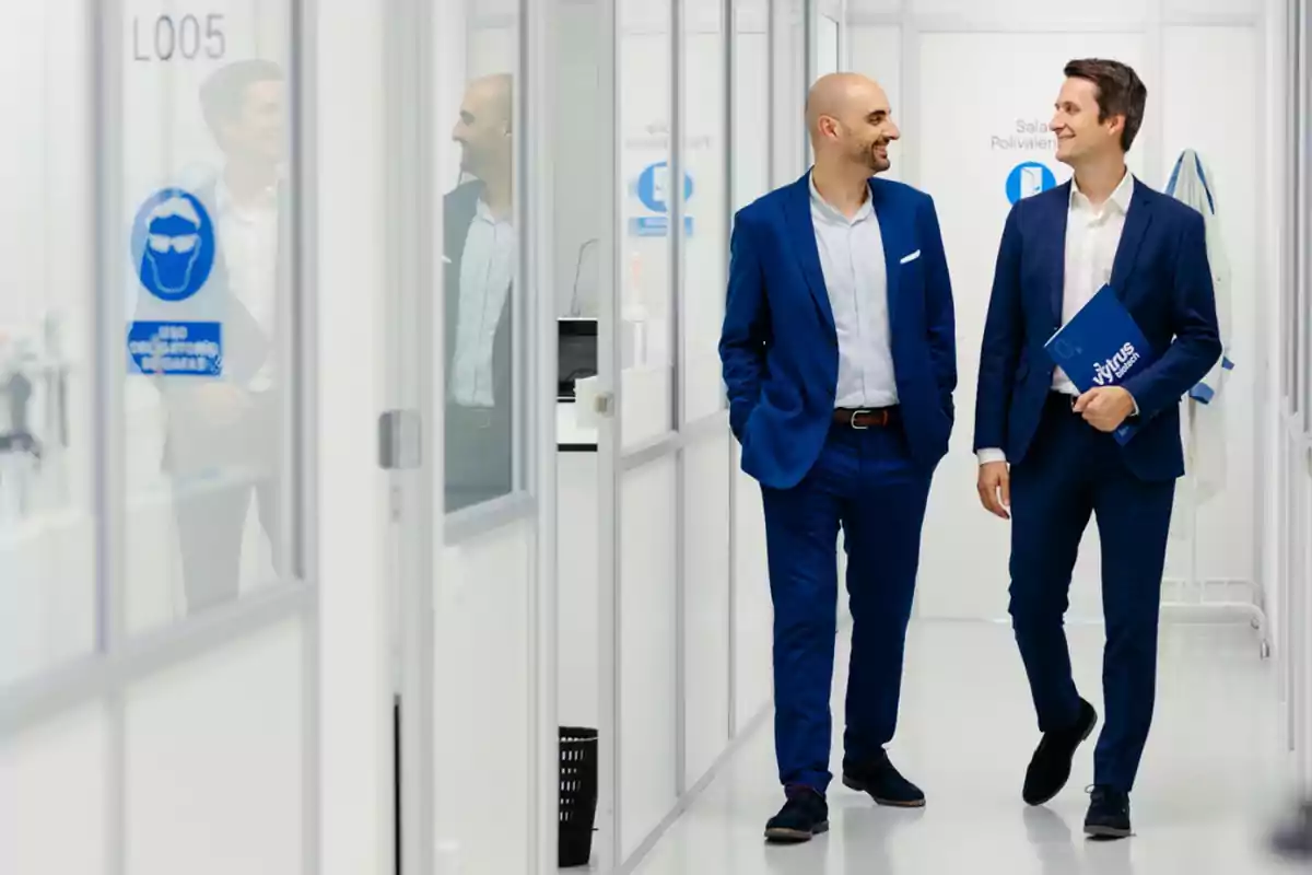 Dos hombres con traje azul caminan y conversan sonrientes por un pasillo moderno y luminoso de oficinas o laboratorio Dos hombres con traje azul caminan y conversan sonrientes por un pasillo moderno y luminoso de oficinas o laboratorio