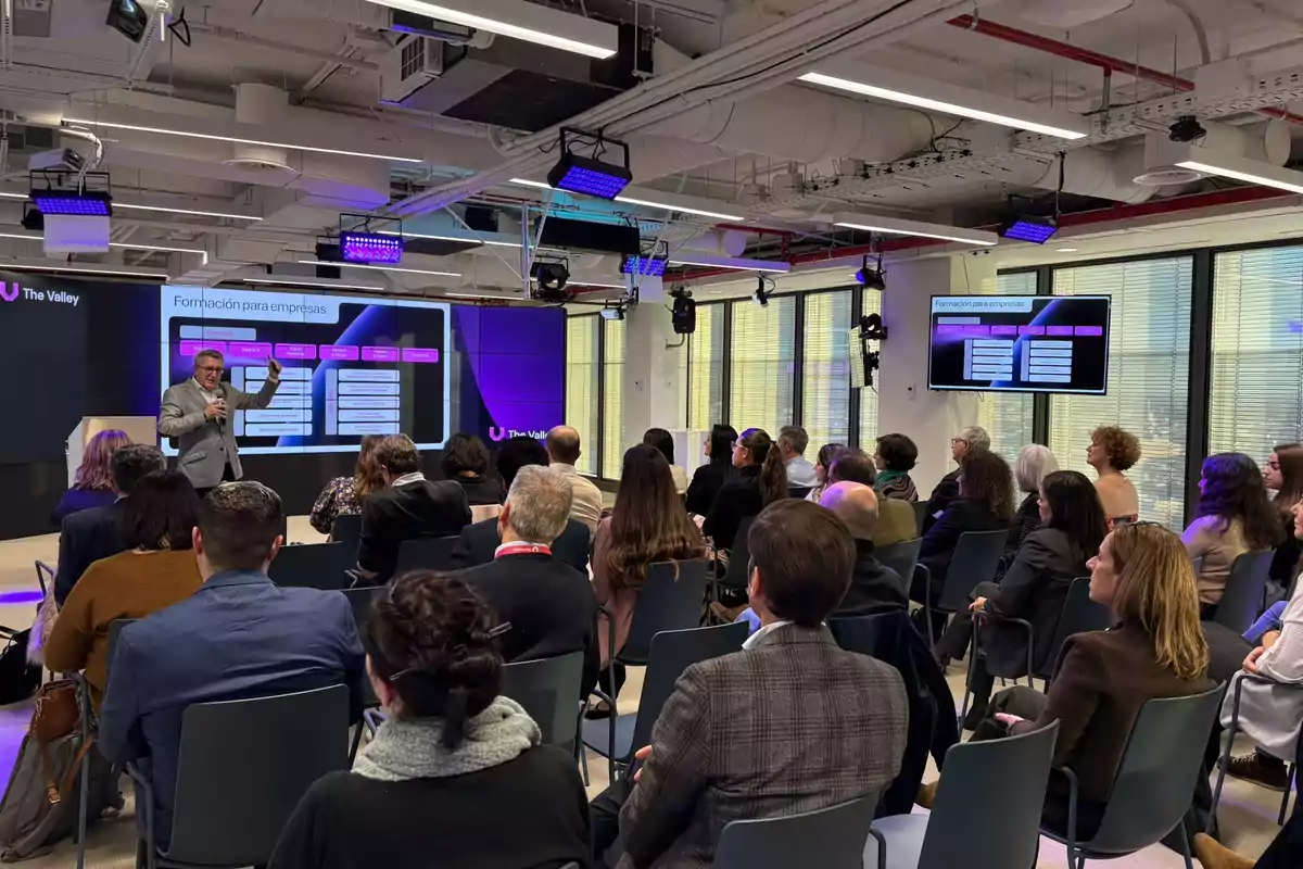 Un grupo de personas asiste a una conferencia en una sala moderna mientras un hombre de pie presenta información en una pantalla grande al frente del auditorio.