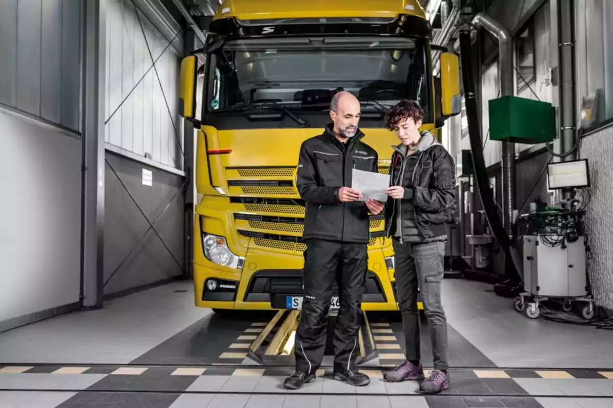 Dos personas revisan documentos frente a un camión amarillo dentro de un taller mecánico Dos personas revisan documentos frente a un camión amarillo dentro de un taller mecánico