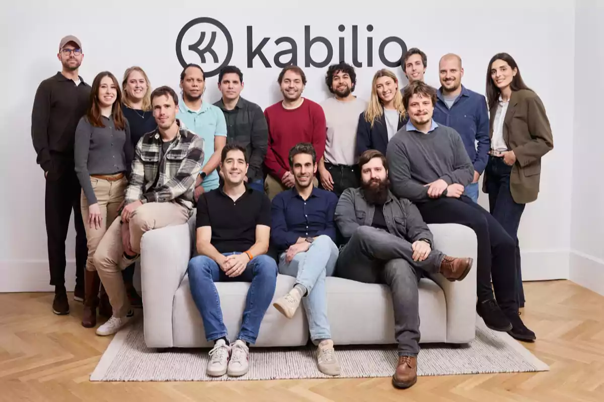 Grupo de personas posando juntas en una oficina frente a una pared con el logo de Kabilio Grupo de personas posando juntas en una oficina frente a una pared con el logo de Kabilio