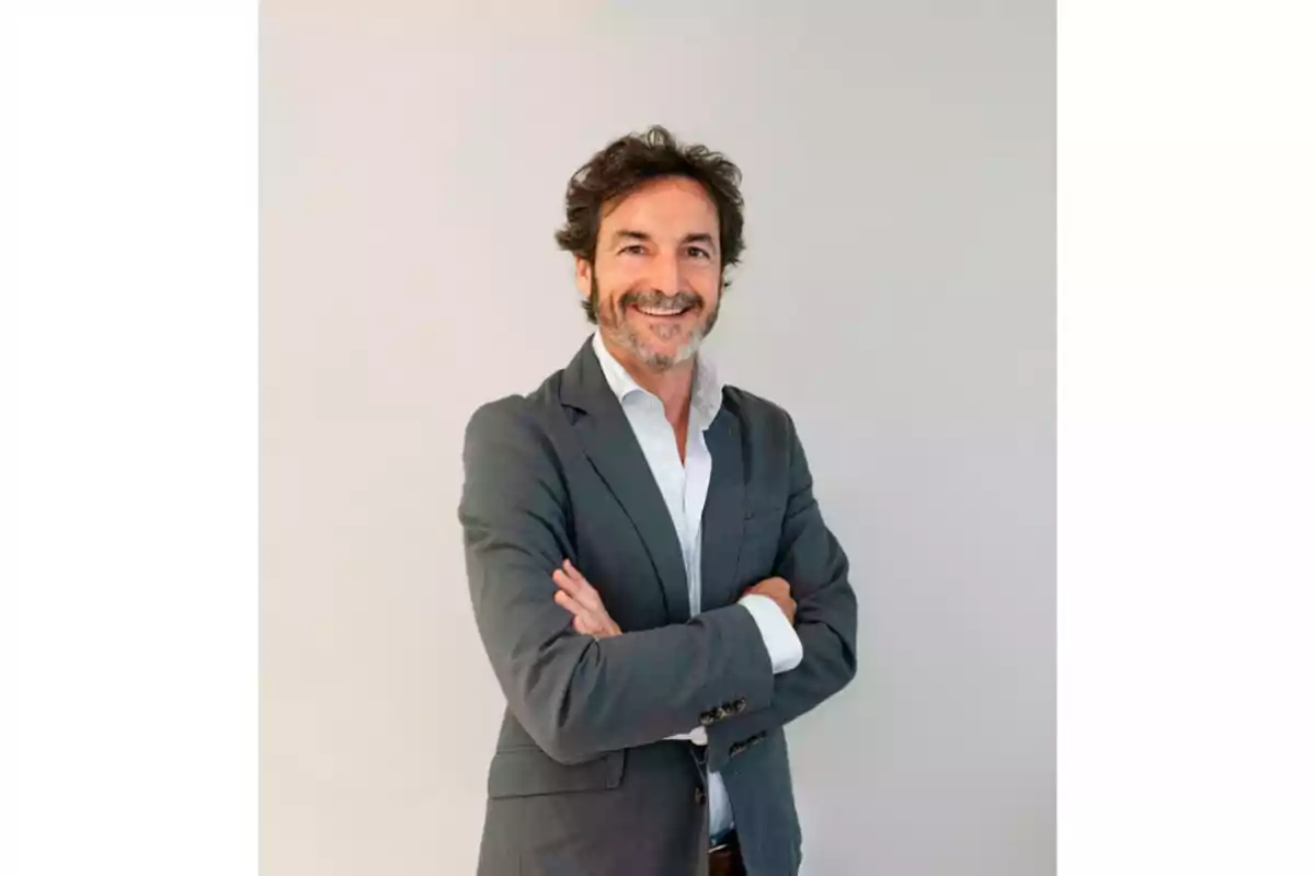 Hombre de mediana edad con barba y cabello oscuro, vestido con saco gris y camisa blanca, sonriendo y con los brazos cruzados frente a un fondo claro