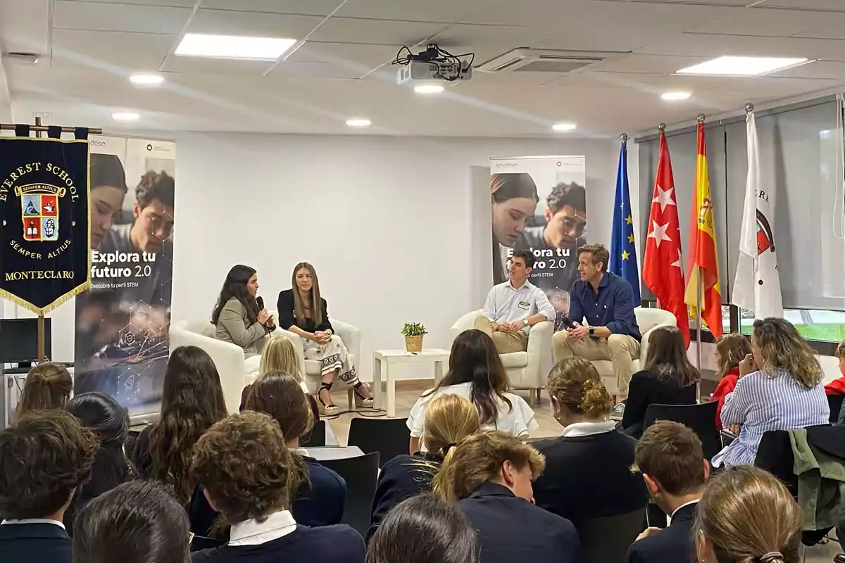 Un grupo de estudiantes asiste a una charla con cuatro ponentes sentados en sillones blancos en un auditorio escolar decorado con banderas y un estandarte de Everest School Montelcarlo