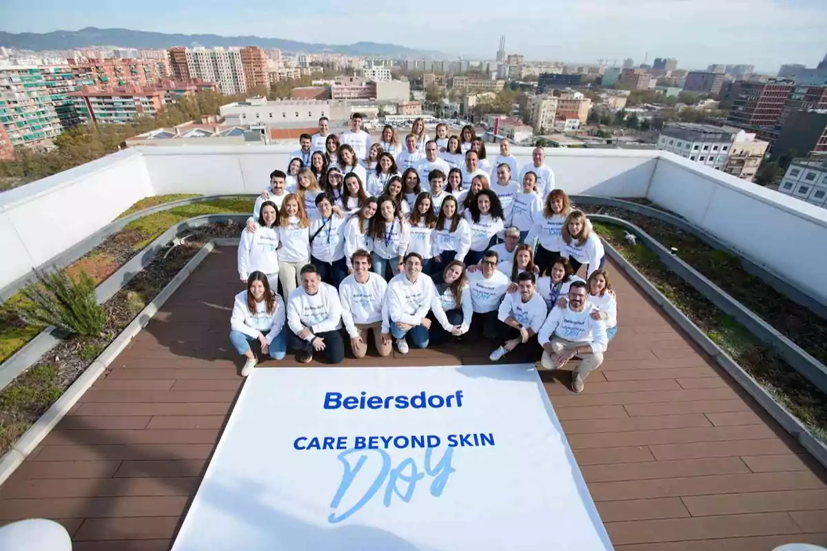 Grupo de personas con sudaderas blancas posando en una azotea junto a un cartel grande que dice Beiersdorf Care Beyond Skin Day con una ciudad al fondo Grupo de personas con sudaderas blancas posando en una azotea junto a un cartel grande que dice Beiersdorf Care Beyond Skin Day con una ciudad al fondo