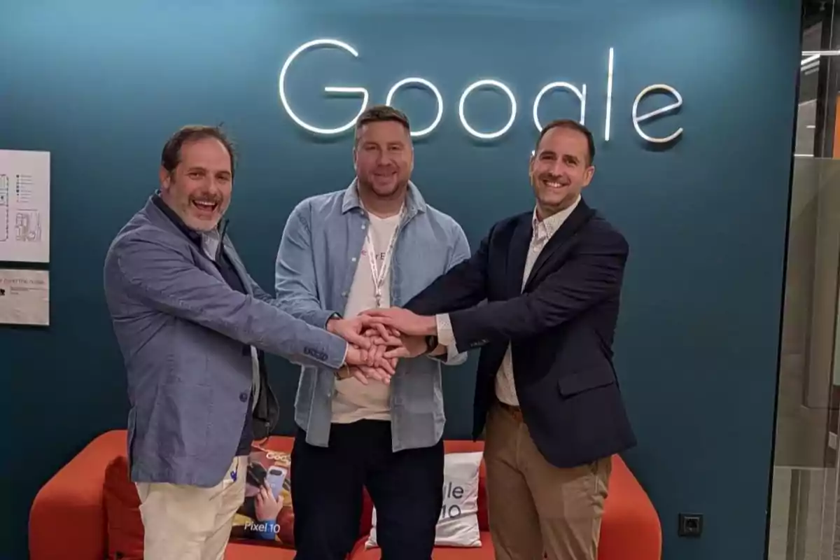 Tres hombres sonrientes se dan la mano frente a una pared azul con el logo de Google iluminado, sentados en un sofá naranja
