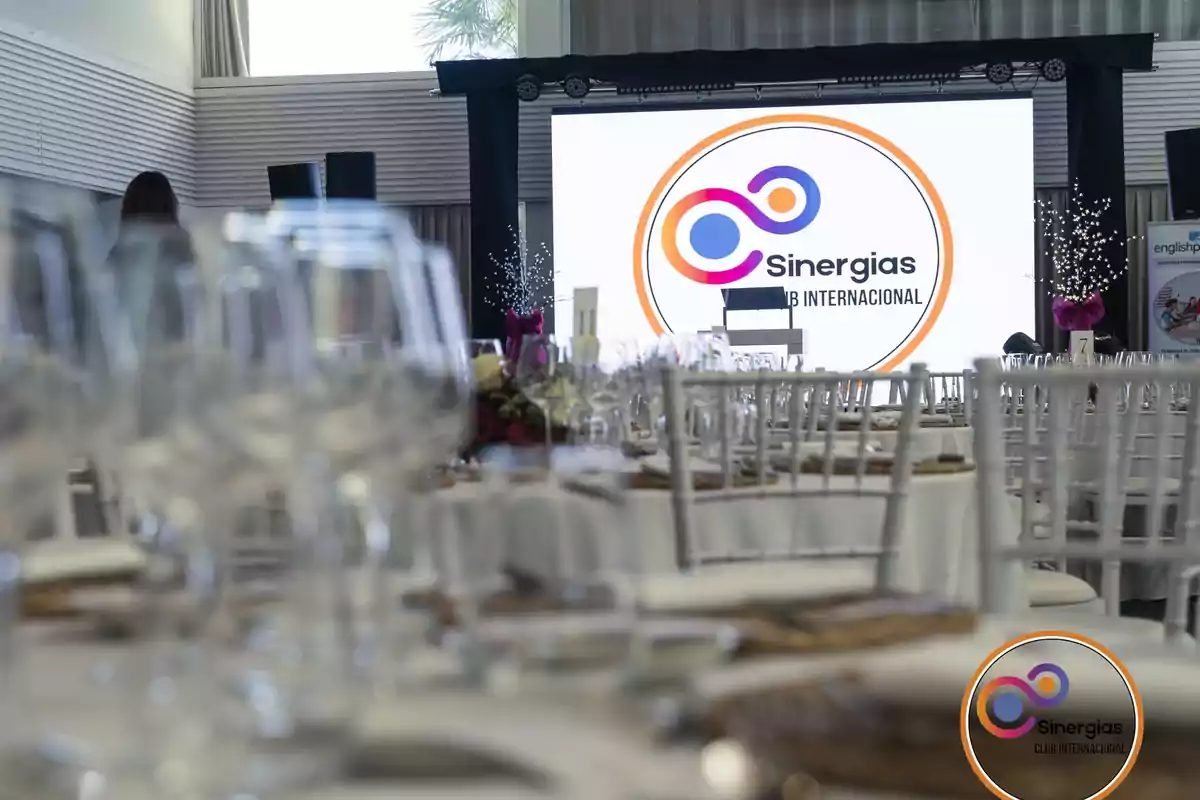 Salón elegantemente decorado con mesas y copas listas para un evento corporativo de Sinergias Club Internacional con un escenario y pantalla al fondo
