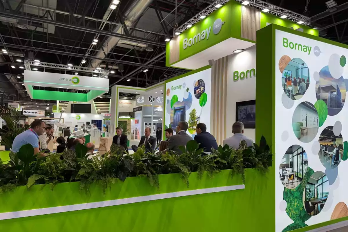 Personas sentadas conversando en un stand de exposición con decoración verde y plantas, donde se observa el nombre Bornay en la estructura y paneles con imágenes circulares de instalaciones y oficinas. Personas sentadas conversando en un stand de exposición con decoración verde y plantas, donde se observa el nombre Bornay en la estructura y paneles con imágenes circulares de instalaciones y oficinas.