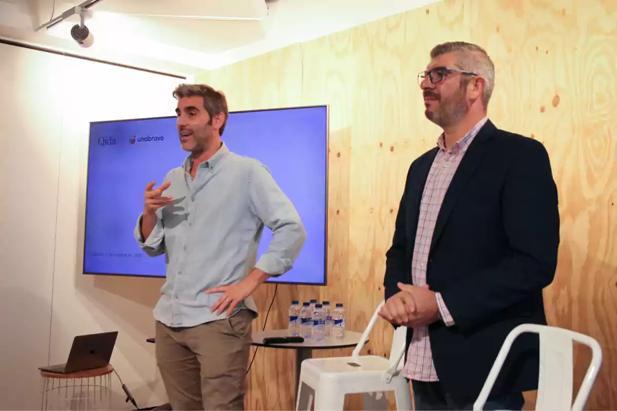 Dos hombres de pie en una sala de conferencias, uno hablando y el otro escuchando, con una pantalla y botellas de agua al fondo Dos hombres de pie en una sala de conferencias, uno hablando y el otro escuchando, con una pantalla y botellas de agua al fondo
